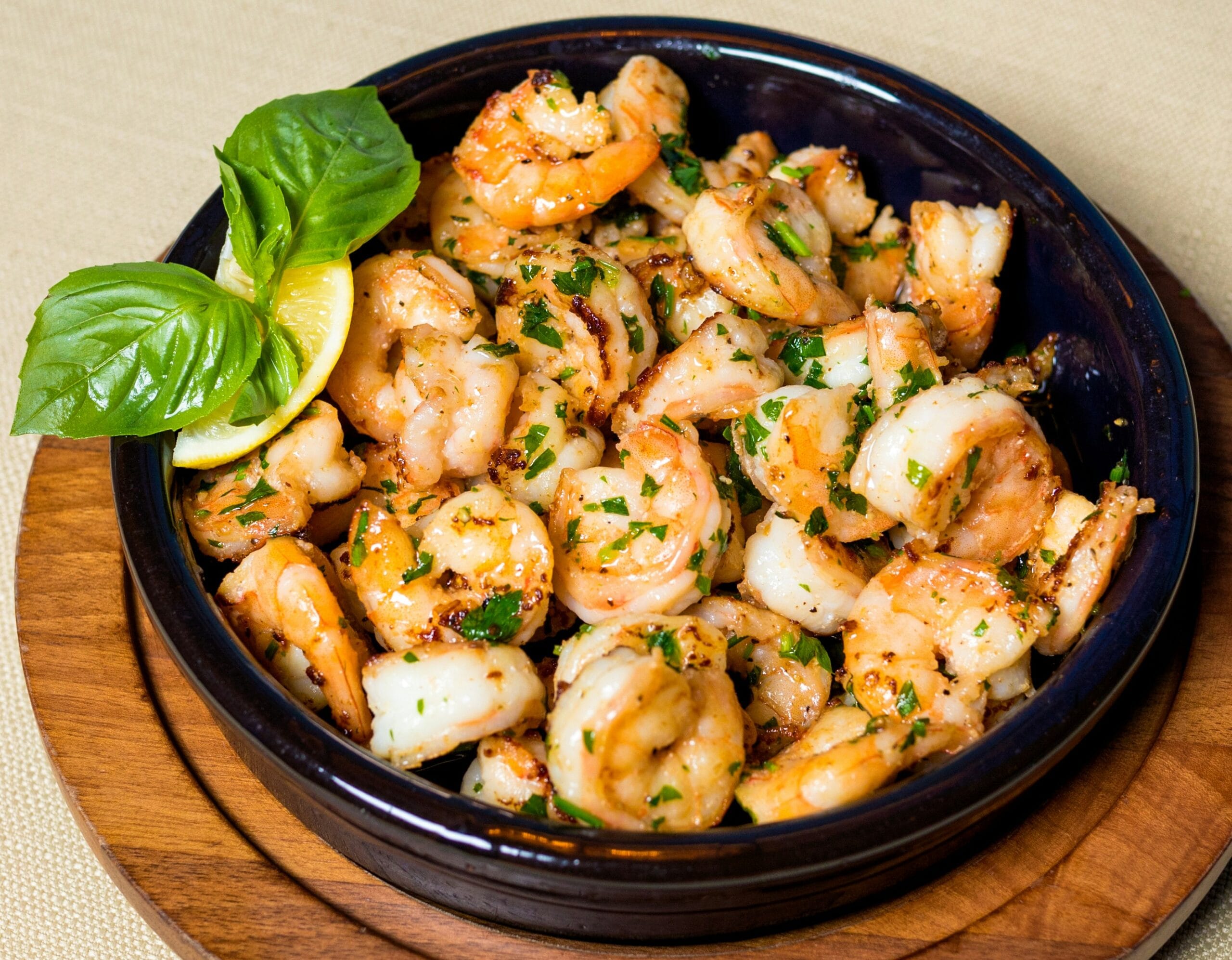 easy Garlic butter shrimp served in a black ceramic dish, garnished with fresh basil leaves and a lemon wedge, on a wooden serving board.