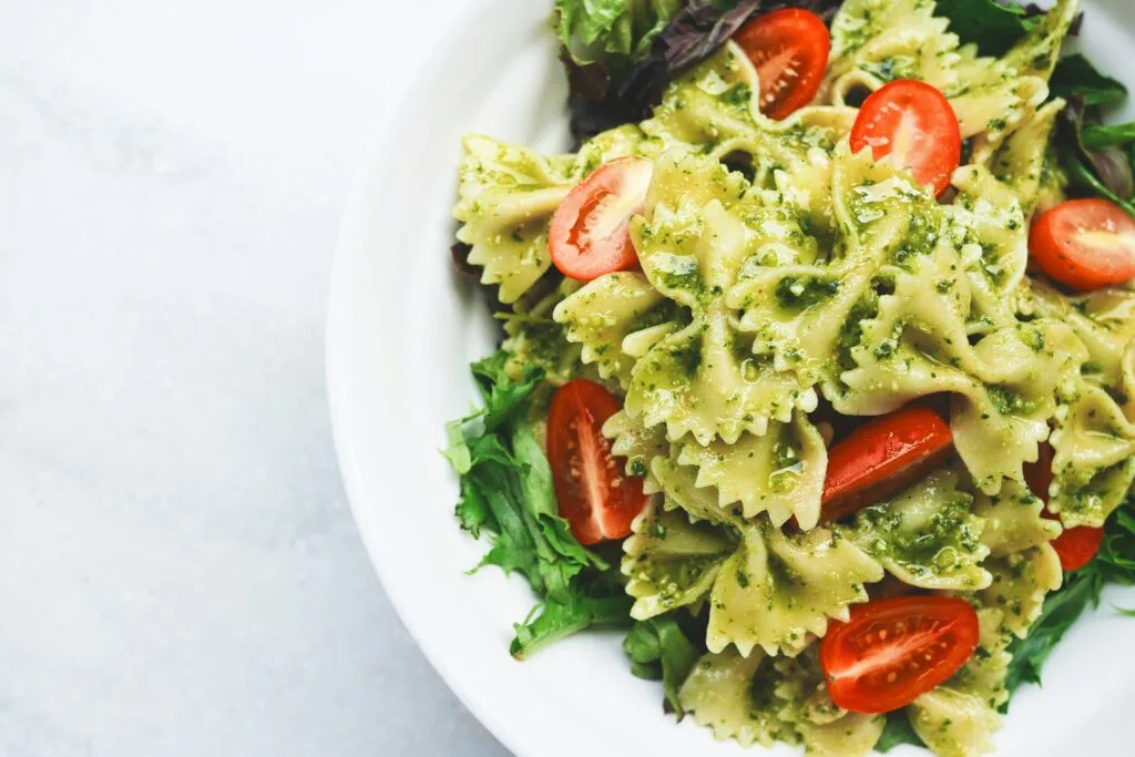 A close-up of a vibrant Healthy Quick Pesto Pasta Salad made with bowtie pasta, cherry tomatoes, and fresh mixed greens, served in a white bowl on a light background.
