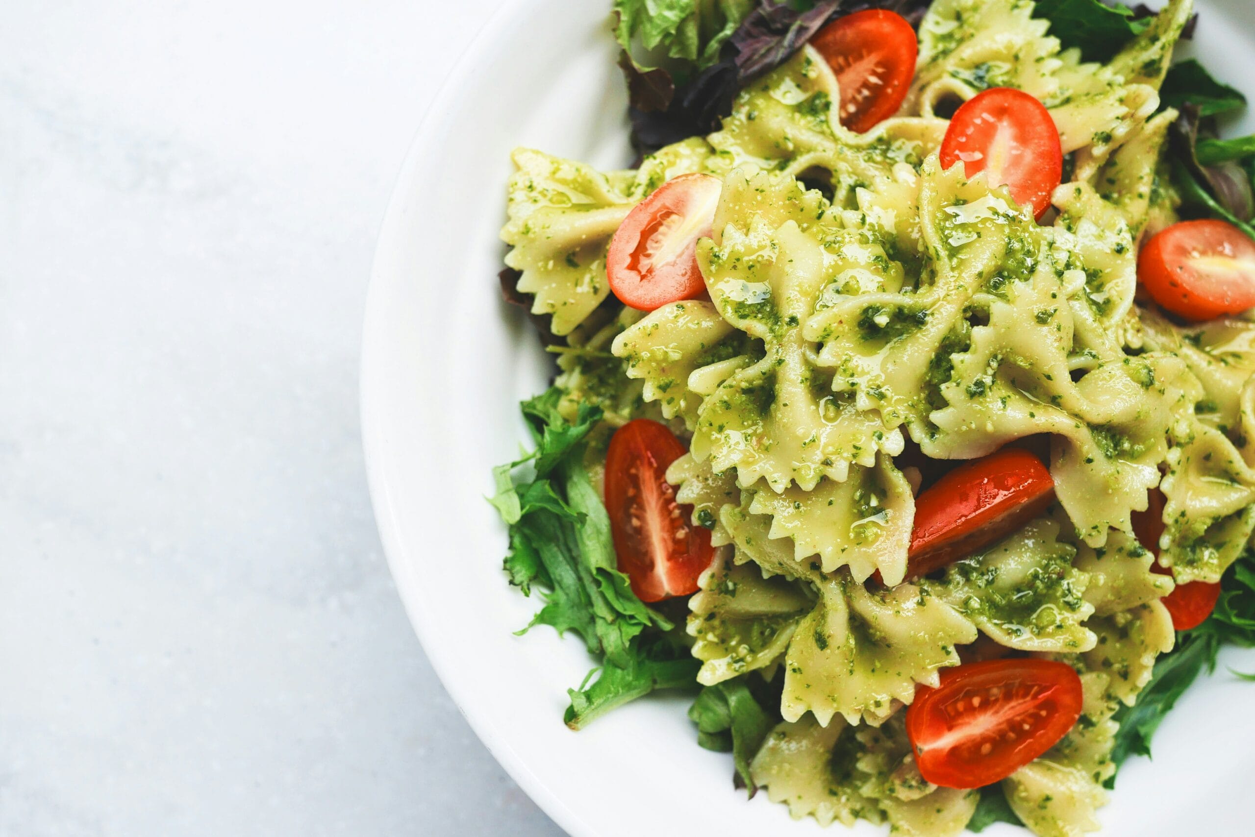 A close-up of a vibrant Healthy Quick Pesto Pasta Salad made with bowtie pasta, cherry tomatoes, and fresh mixed greens, served in a white bowl on a light background.