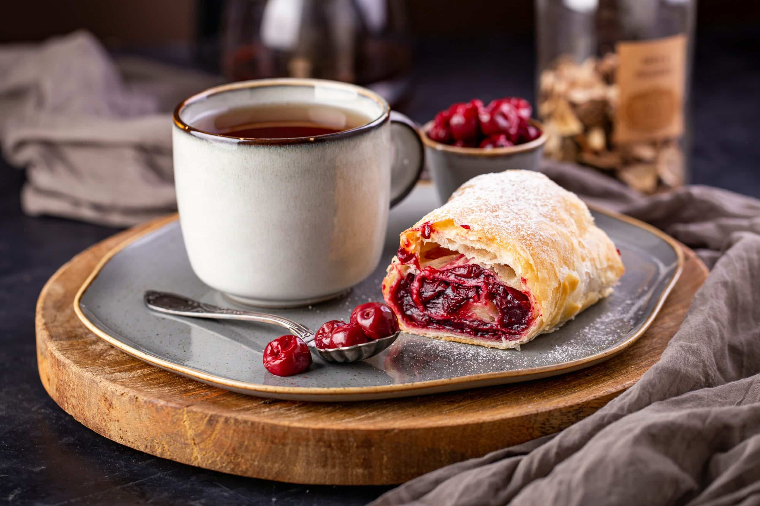 A golden, flaky cherry strudel with a dusting of powdered sugar, served alongside a warm cup of tea—an inviting breakfast pastries.