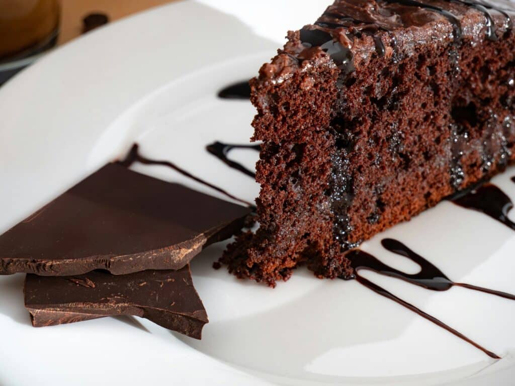 A well-lit kitchen countertop displaying an assortment of healthy chocolate cake ingredients, including organic cocoa powder, oat flour, ripe avocados, melted dark chocolate, a bowl of honey, and a carton of Greek yogurt. In the foreground, a wooden cutting board holds chopped walnuts and shredded unsweetened coconut. The lighting is soft and natural, casting warm shadows that accentuate the rich colors of the ingredients. The overall composition is clean, minimalist, and inviting, conveying a sense of wholesome indulgence. Healthy Chocolate Cake
